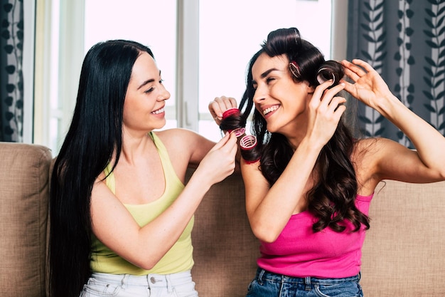 Two young beautiful smiling girl friends sitting on the couch at home make each other hairstyles and communicate fun