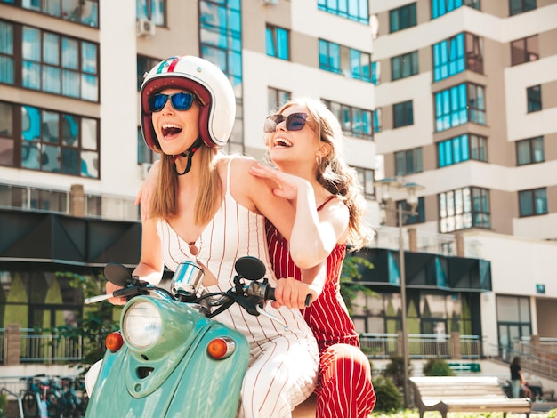 Two young beautiful smiling hipster female in trendy overallsSexy carefree women driving retro motorbike on the street backgroundPositive models having fun riding classic Italian scooter in eyewear