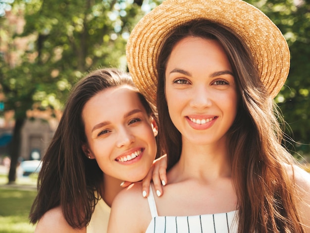 Two young beautiful smiling hipster female in trendy summer clothesSexy carefree women posing on the street background in hat Positive pure models having fun at sunset hugging