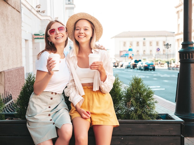 Two young beautiful smiling hipster female in trendy summer clothesSexy carefree women posing in the street Positive pure models having fun at sunset They drinking coffee or tea in plastic cup