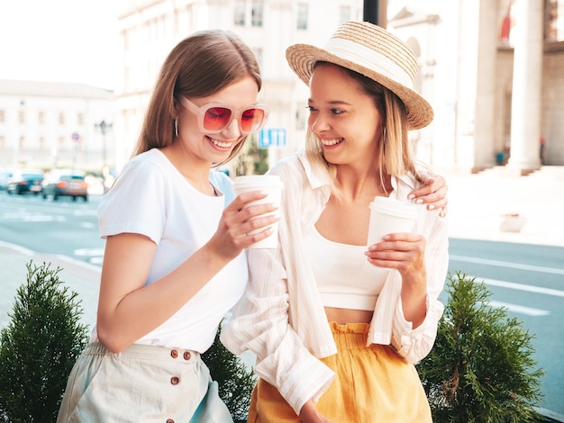 Two young beautiful smiling hipster female in trendy summer clothesSexy carefree women posing in the street Positive pure models having fun at sunset They drinking coffee or tea in plastic cup