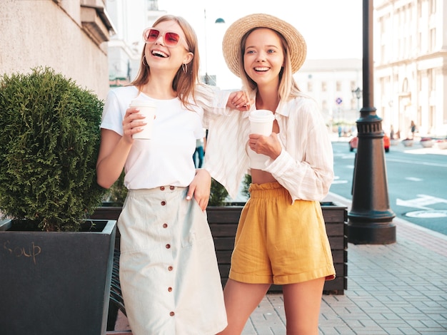 Two young beautiful smiling hipster female in trendy summer clothesSexy carefree women posing in the street Positive pure models having fun at sunset They drinking coffee or tea in plastic cup