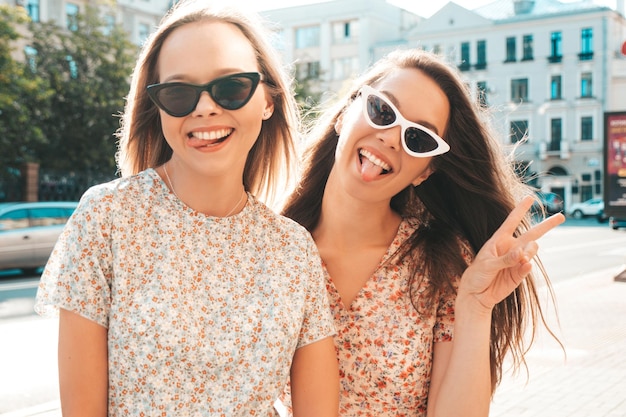 Two young beautiful smiling hipster female in trendy summer dressesSexy carefree women posing in the street Positive pure models having fun at sunset hugging and going crazy Show peace sign