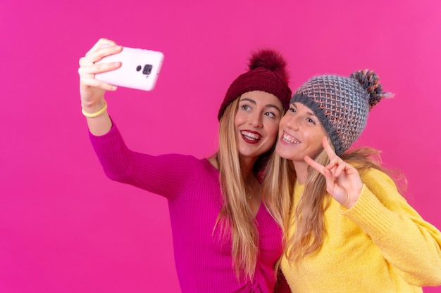 Two young blonde caucasian women with a phone isolated on a pink background selfie photo smiling