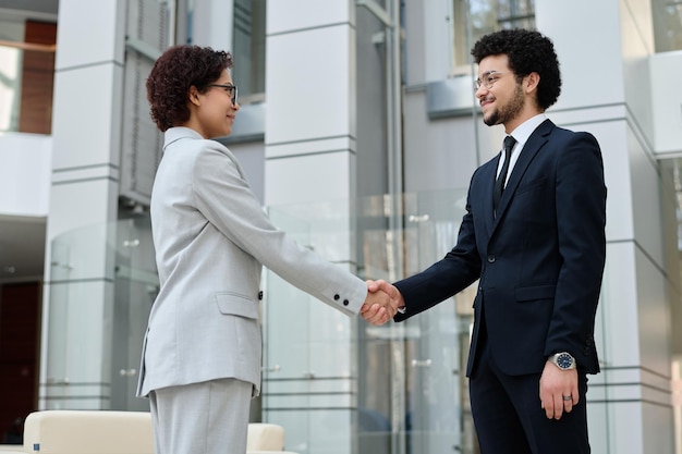 Two young business colleagues shaking hands while greeting each other standing in office hall