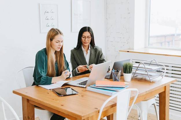 Two young business women professionals in formal wear clothes work in modern office using laptop tablet brainstorm and search for solutions together confident independent Asian girl solves problems