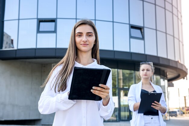 Two young business women with tablet and clipboard posing outside business center