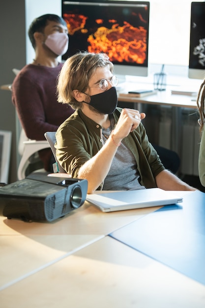 Two young businessmen in casualwear and protective masks sitting by desks and listening to coach while looking at whiteboard at seminar