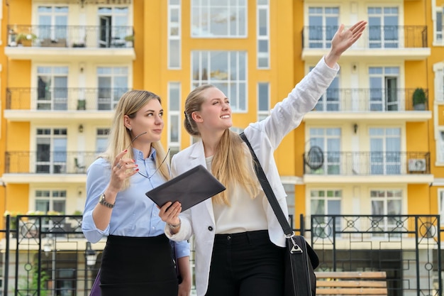 Two young businesswomen on city street talking and looking in screen of digital tablet