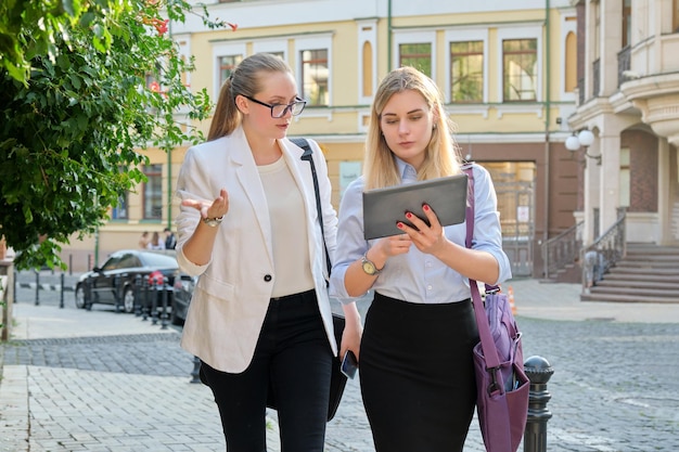 Two young businesswomen walking along city street, talking and looking in screen of digital tablet. Business, people, technology, teamwork, communication concept