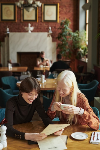 Two young cheerful women in casualwear reading menu paper in cafe