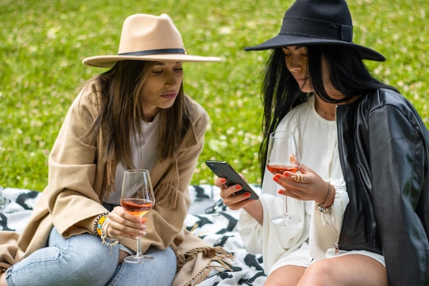 Two young cheerful women sitting on a blanket in the park looking an the phone and drinking wine while on a picnic