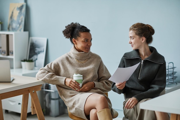 Two young colleagues discussing project and drinking coffee together during break at office