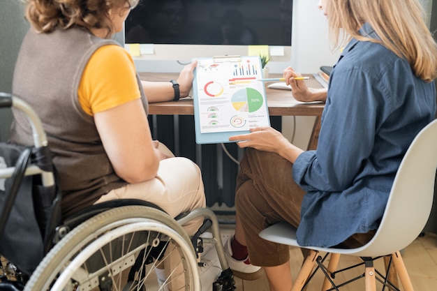 Two young contemporary female economists discussing financial papers with charts and diagrams while one of them explaining data