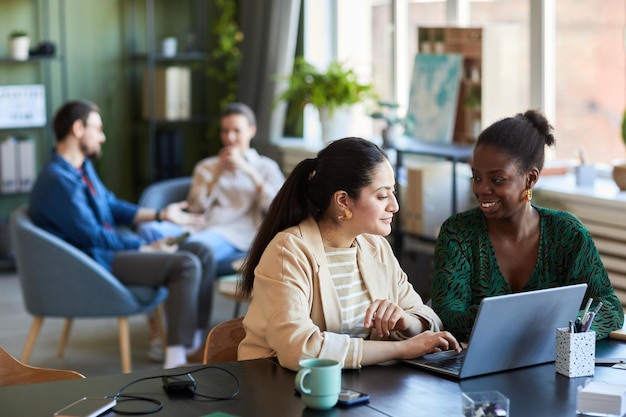 Two young creative employees discussing ideas while sitting by desk in front of laptop and working o