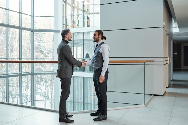 Two young elegant business partners shaking hands of each other while making deal inside large office building