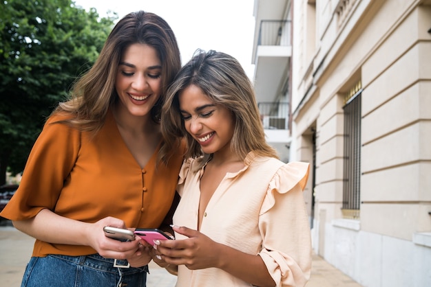 Two young friends enjoying together and using their mobile phones while standing outdoors on the street