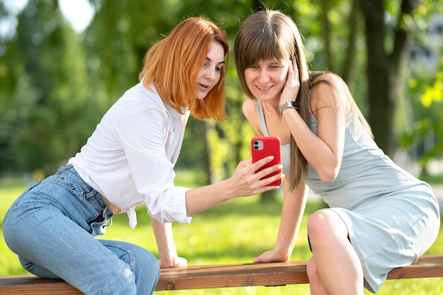 Two young girls friends sitting on a bench in summer park looking in red smartphone.