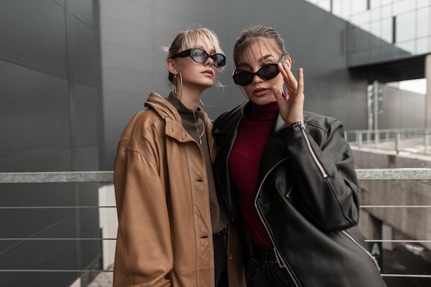 Two young girls sisters with vogue sunglasses in a fashionable leather jacket and stylish knitter sweaters posing on the street near a metal wall