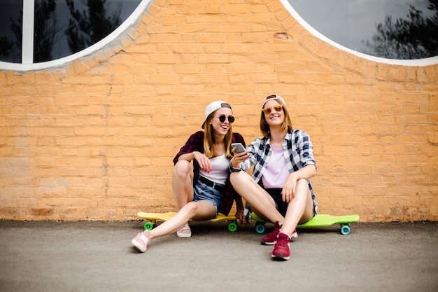 Two young happy girl friends in hipster outfit sitting on longboards and making selfie on phone.