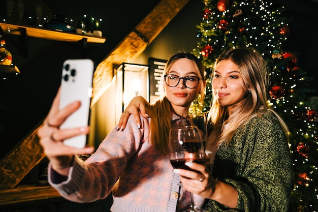 Two young happy women making festive photo selfie near Christmas tree and drinking wine.