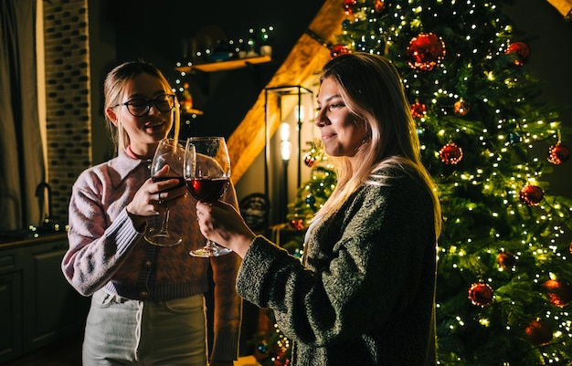 Two young happy women toasting and drinking wine near Christmas tree.