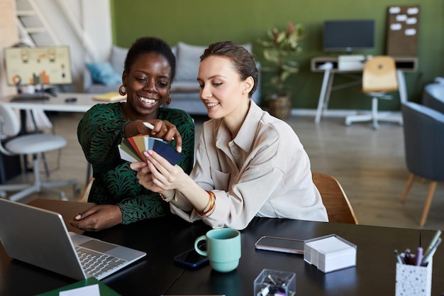 Two young intercultural businesswomen choosing color from palette while working over creating new pr