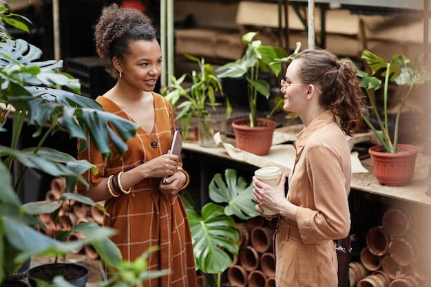 Two young intercultural female florists in smart casualwear having discussion against shelf with plants in pots in orangerie or flower shop