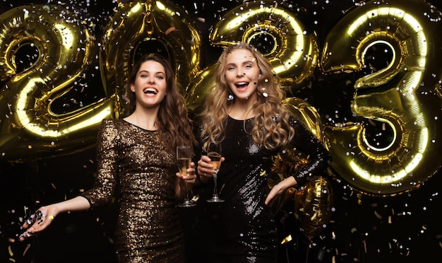 Two young ladies drinking champagne Image of girls with balloons isolated on black background having fun at New Year's 2023 Eve Party