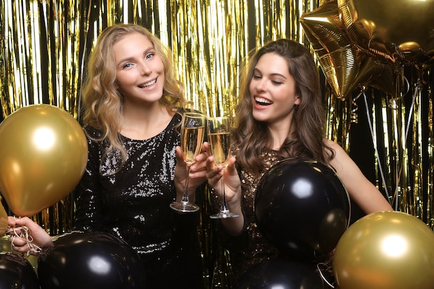 Two young ladies drinking champagne. Image of girls with balloons isolated on gold background.