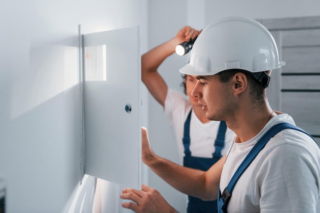 Two young male electricians works indoors together Using flashlight