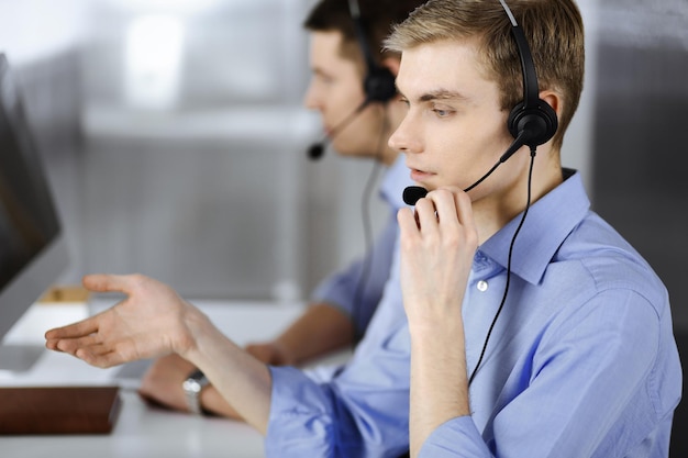 Two young men in headset, sitting at the desk in the modern office, listening to the clients. Call center operators at work.