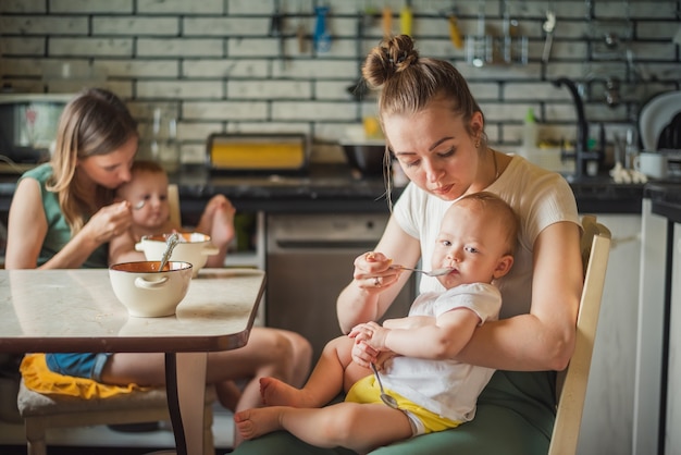 Two young mothers together feed their happy babies milk porridge in the kitchen