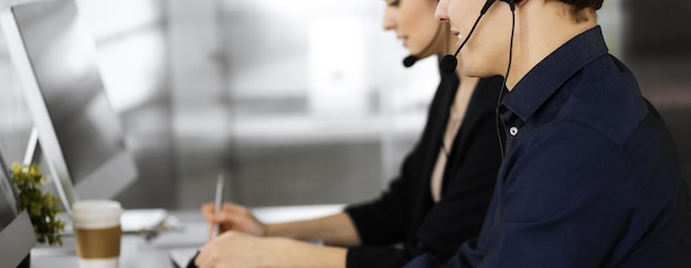 Two young people in headsets are talking to the clients, while sitting at the desk in a modern office. Focus on man in a blue shirt. Call center operators at work.