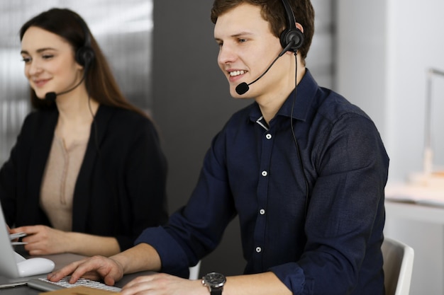 Two young people in headsets are talking to the clients, while sitting at the desk in a modern office. Focus on man in a blue shirt. Call center operators at work.