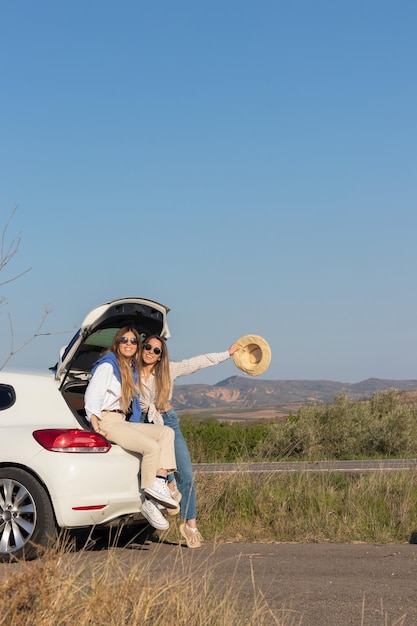 Two young pretty lady sitting in an open car boot having fun enjoying their summer road trip through countryside