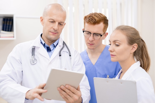 Two young serious interns listening attentively to confident clinician while looking at information on touchpad screen