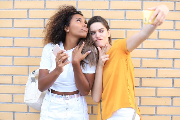 Two young smiling hipster women in summer clothes posing on street.Female showing positive face emotions.