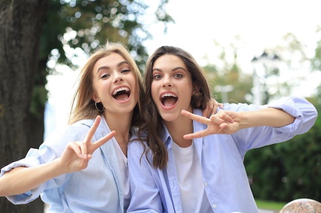 Two young smiling hipster women in summer clothes posing on street.Female showing positive face emotions.