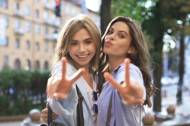 Two young smiling hipster women in summer clothes posing on street.Female showing positive face emotions.