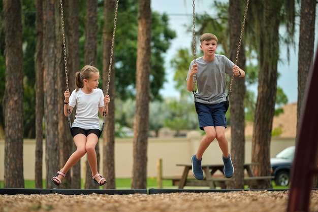 Two young teenage children girl and boy playing on swings together outdoors on bright sunny vacations day