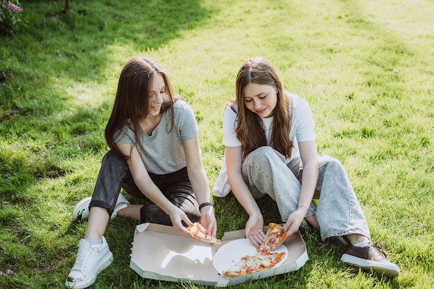 Two young teenage friends having fun in the park on the grass and eating pizza. Women eat fast food. Not a healthy diet. Soft selective focus.