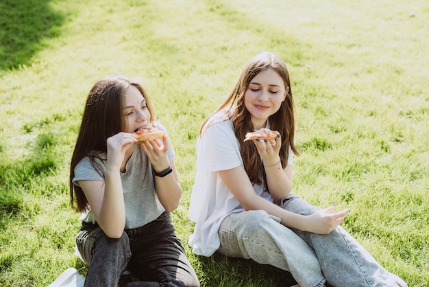 Two young teenage friends having fun in the park on the grass and eating pizza. Women eat fast food. Not a healthy diet. Soft selective focus.