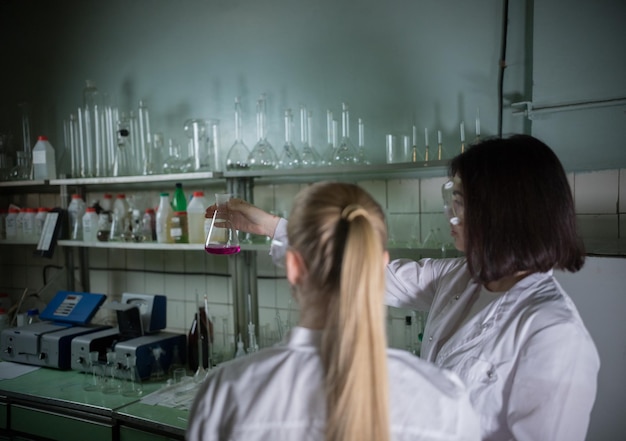 Two young woman in chemical laboratory holding a flask with pink liquid in it back view