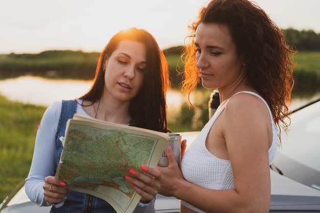 Two young women are traveling on the roads by car stopped at the side of the road and look at the map drinking coffee from a thermos Vacation concept