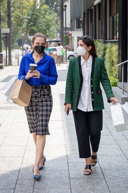 Two young women are walking down the street wearing face masks talking happily after shopping