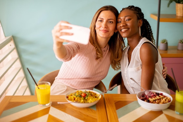 Two young women caucasian and black one taking selfie with mobile phone in the cafe