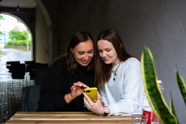 Two young women communicate and look into smartphone in cafe on terrace