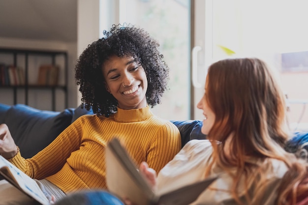 Two young women at home a couple of lesbian sitting in sofa in living room reading books and having moments of relax smiling african woman looking her girlfriend