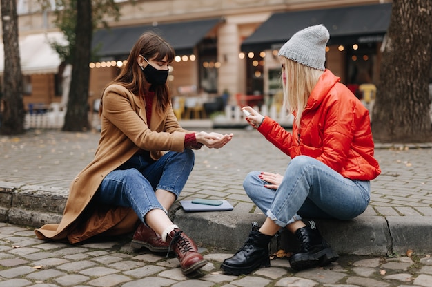 Two young women in medical masks spending free time together on fresh air and using antiseptic for hands. Prevention of coronavirus spreading.